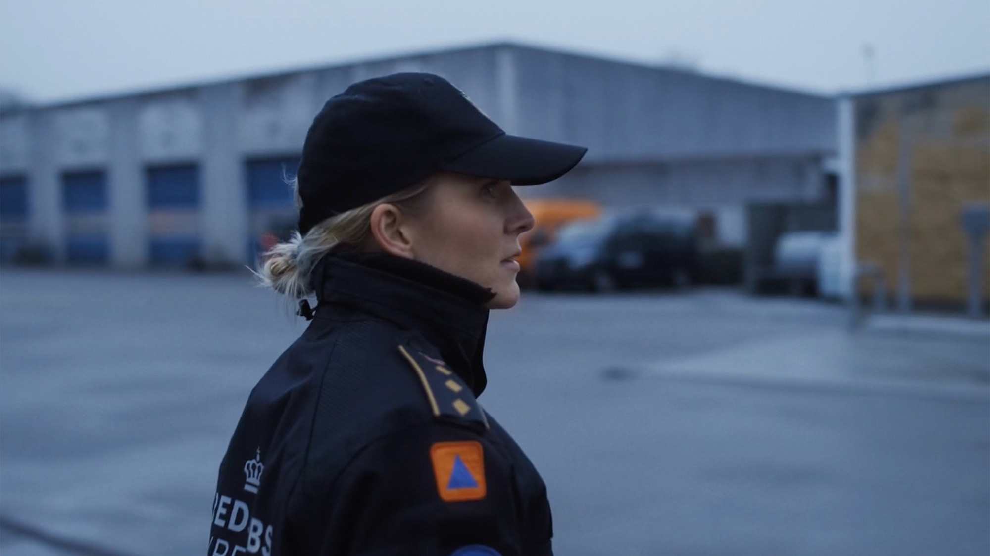 En person i uniform og kasket står udendørs på et gråt område med bygninger og parkerede biler i baggrunden.