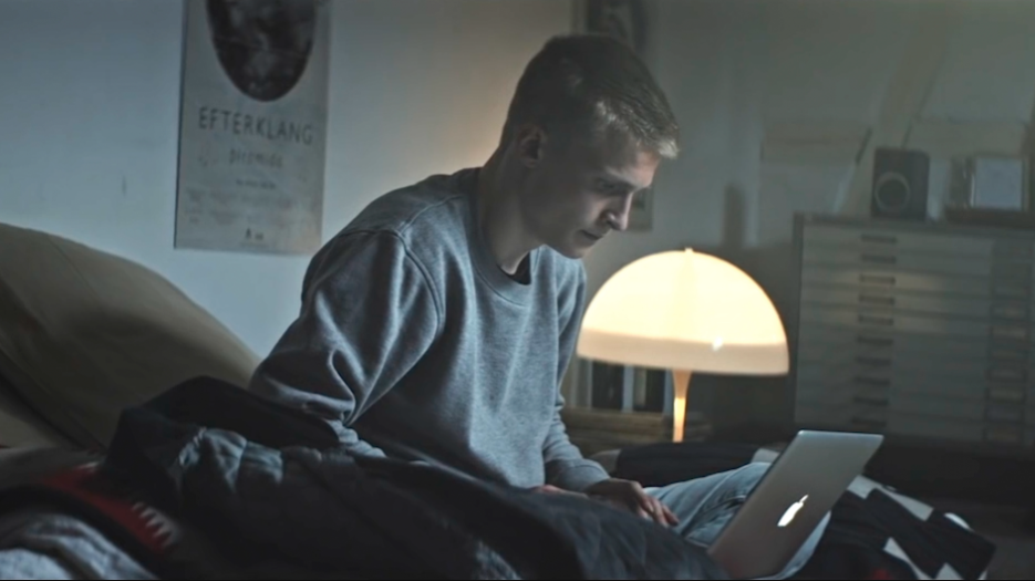 En ung person sidder på en seng i et dæmpet oplyst værelse og arbejder på en bærbar computer.