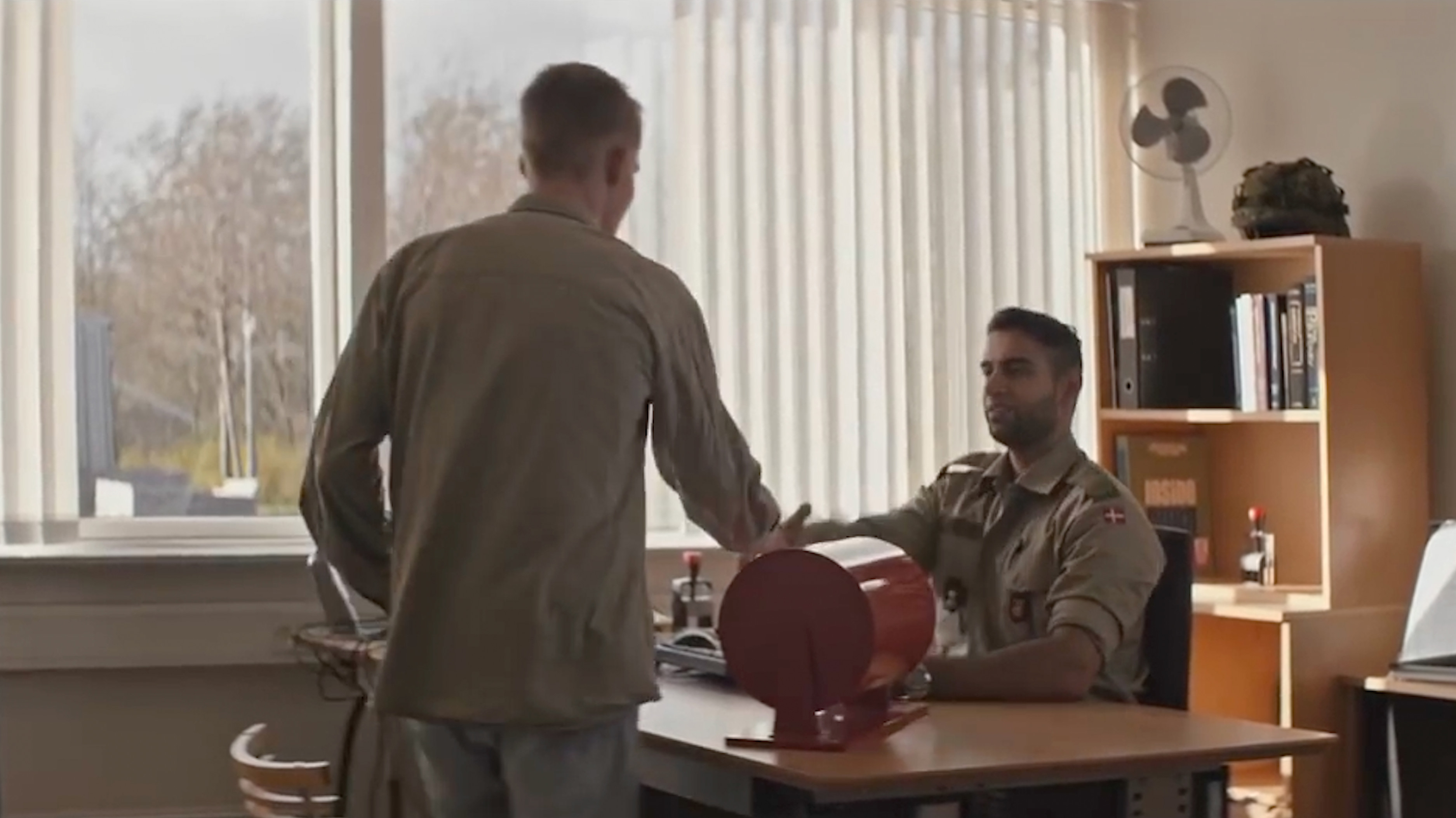 En person i uniform sidder ved et skrivebord og giver hånd til en besøgende, der står foran ham i et kontor med store vinduer og reoler i baggrunden.