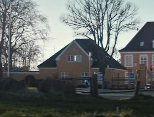 Indgangen til en kaserne med gule bygninger, træer uden blade og en vej i forgrunden på en klar dag.