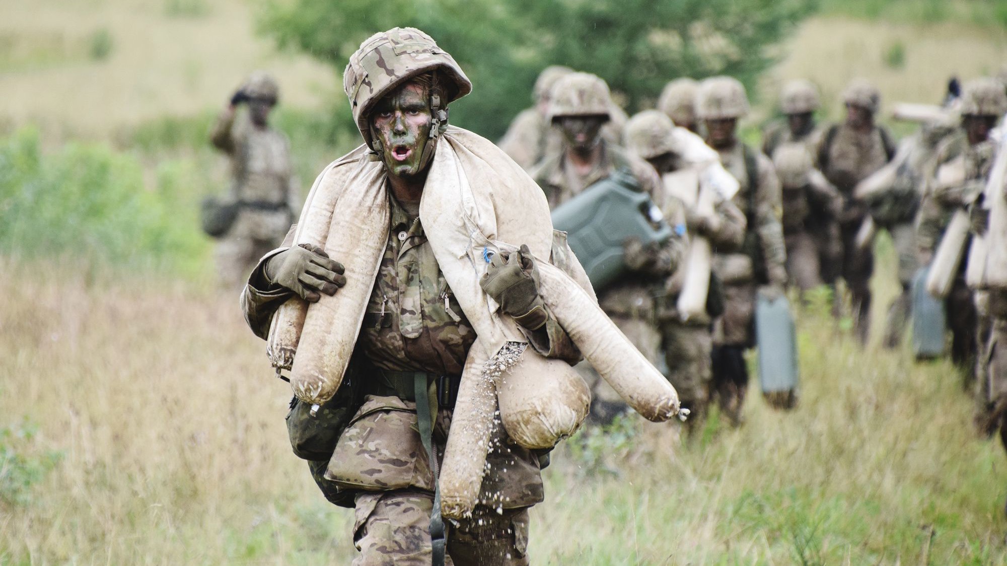 En soldat bærer sandsække under en øvelse, mens en enhed følger efter i baggrunden.