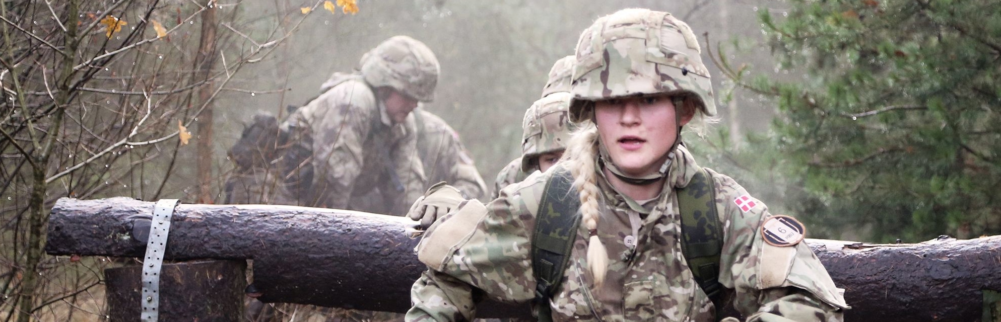 Kvindelig soldat i camouflage klarer en træstamme på en forhindringsbane, mens holdkammerater følger efter i skoven.