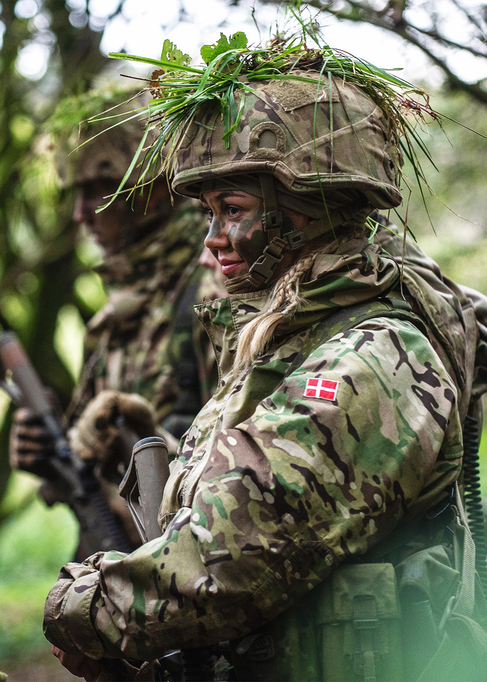 Kvindelig værnepligtig i camouflage og ansigtsmaling står i skoven under en øvelse.