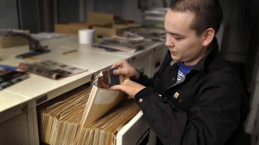 En person står ved et arkivskab og bladrer i en mappe med dokumenter i et kontormiljø.
