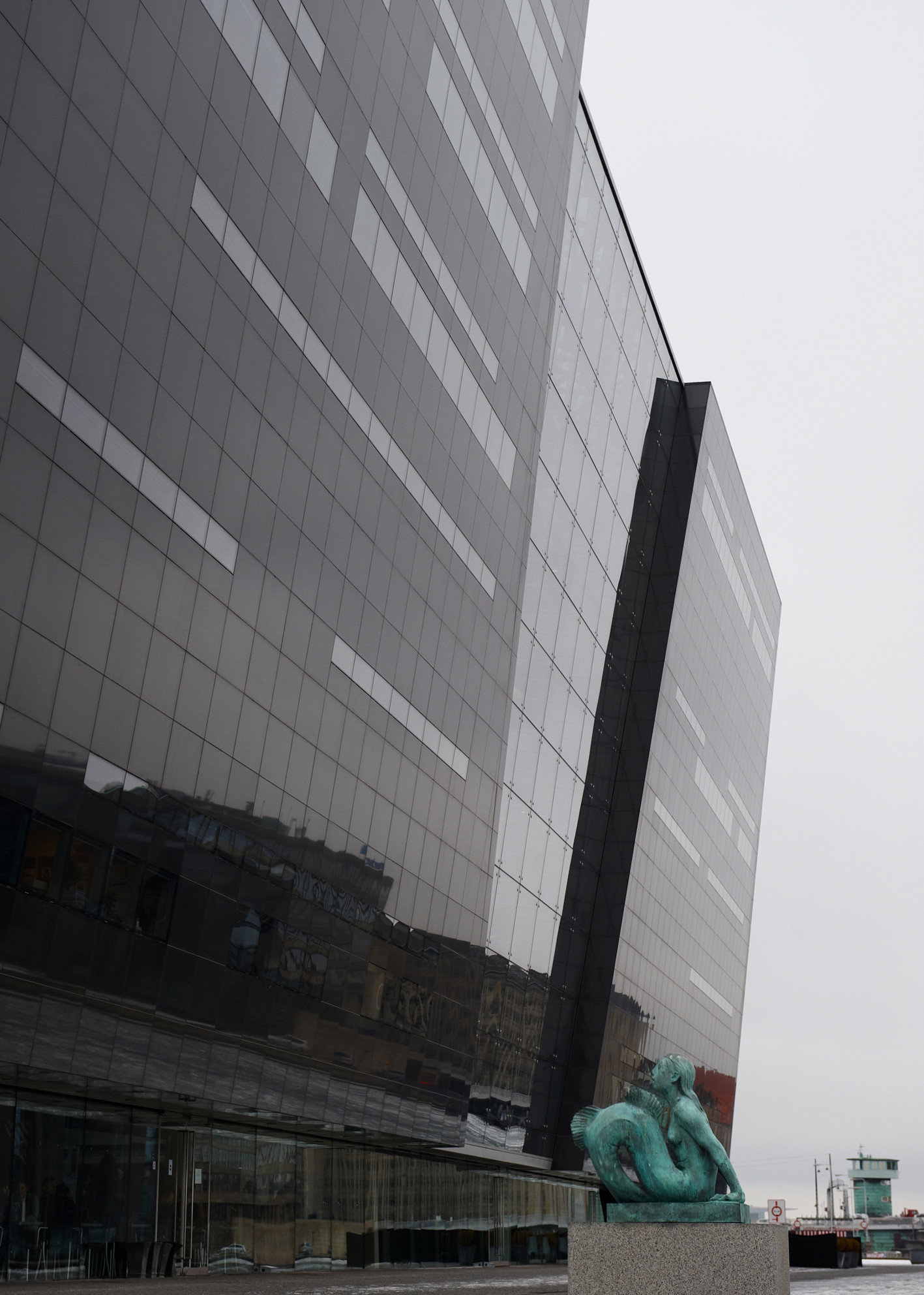tEn moderne glasbygning med mørke facader og en grøn bronzeskulptur foran på en grå og overskyet dag.