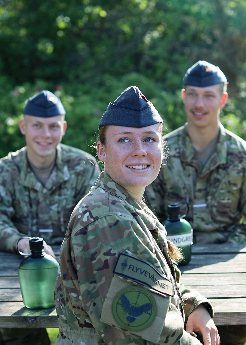 Tre personer i militæruniform sidder ved et udendørs bord i sollys. Personen forrest smiler mod kameraet, mens de to andre sidder bagved og ser i samme retning.