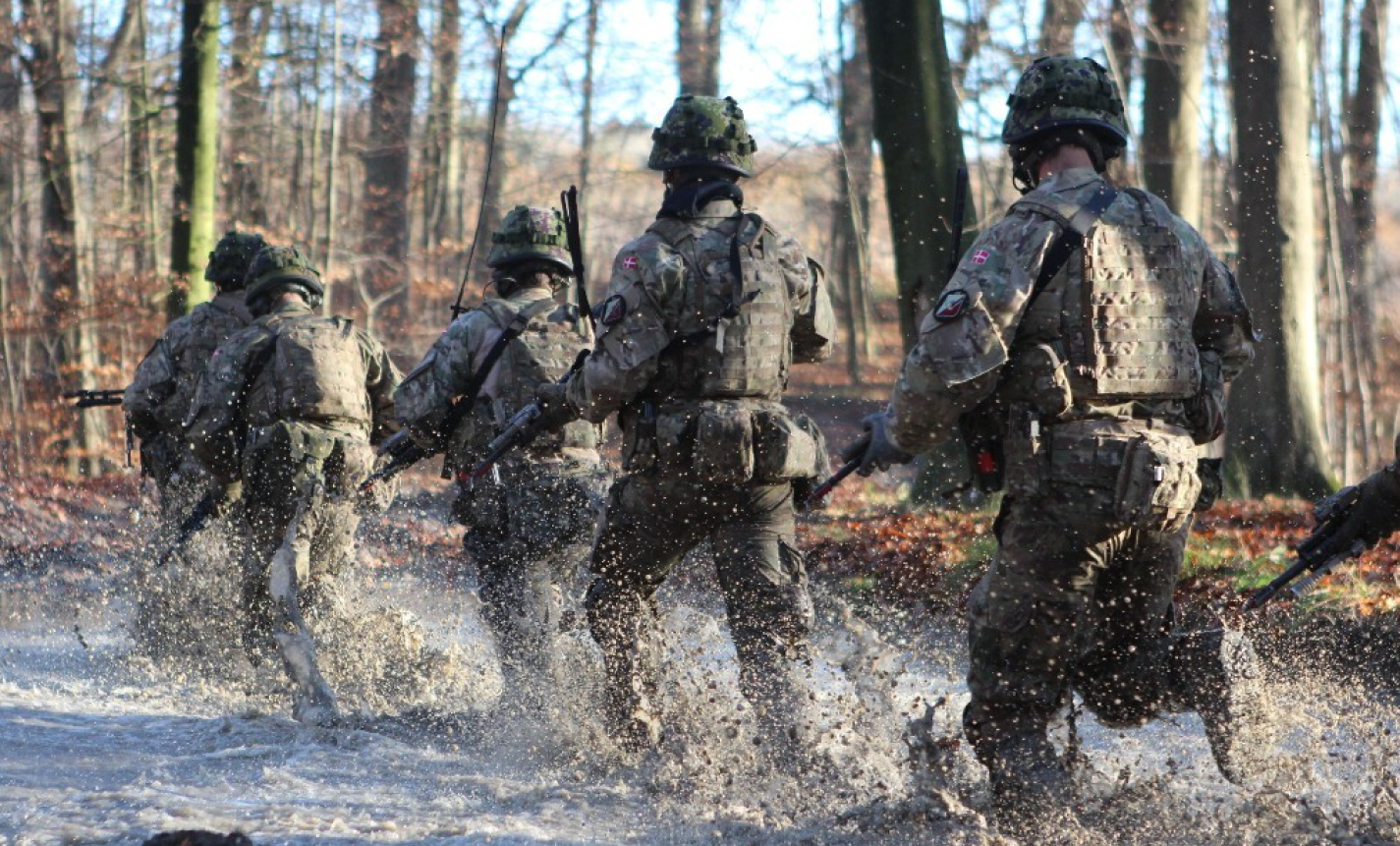 En gruppe soldater i uniform løber gennem lavt vand i en skov, så vandet sprøjter omkring dem, mens de bevæger sig fremad med deres udrustning og våben.