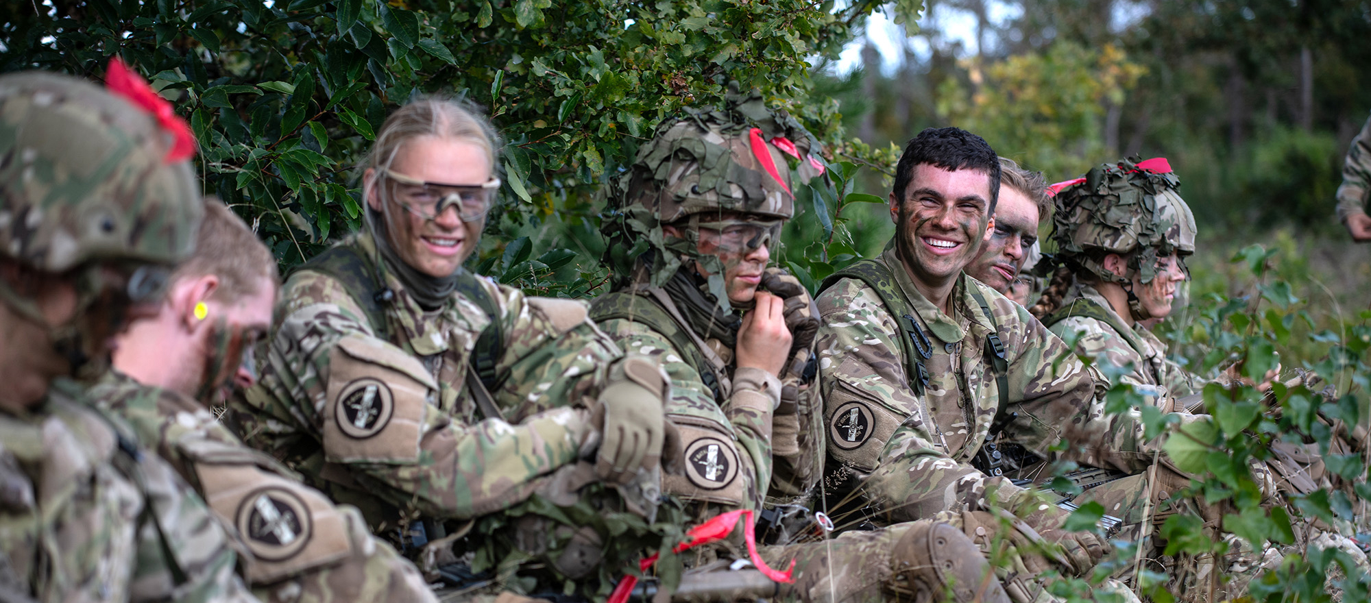 Værnepligtige i camouflageuniformer sidder i terrænet og holder pause med smil og kammeratlig stemning.