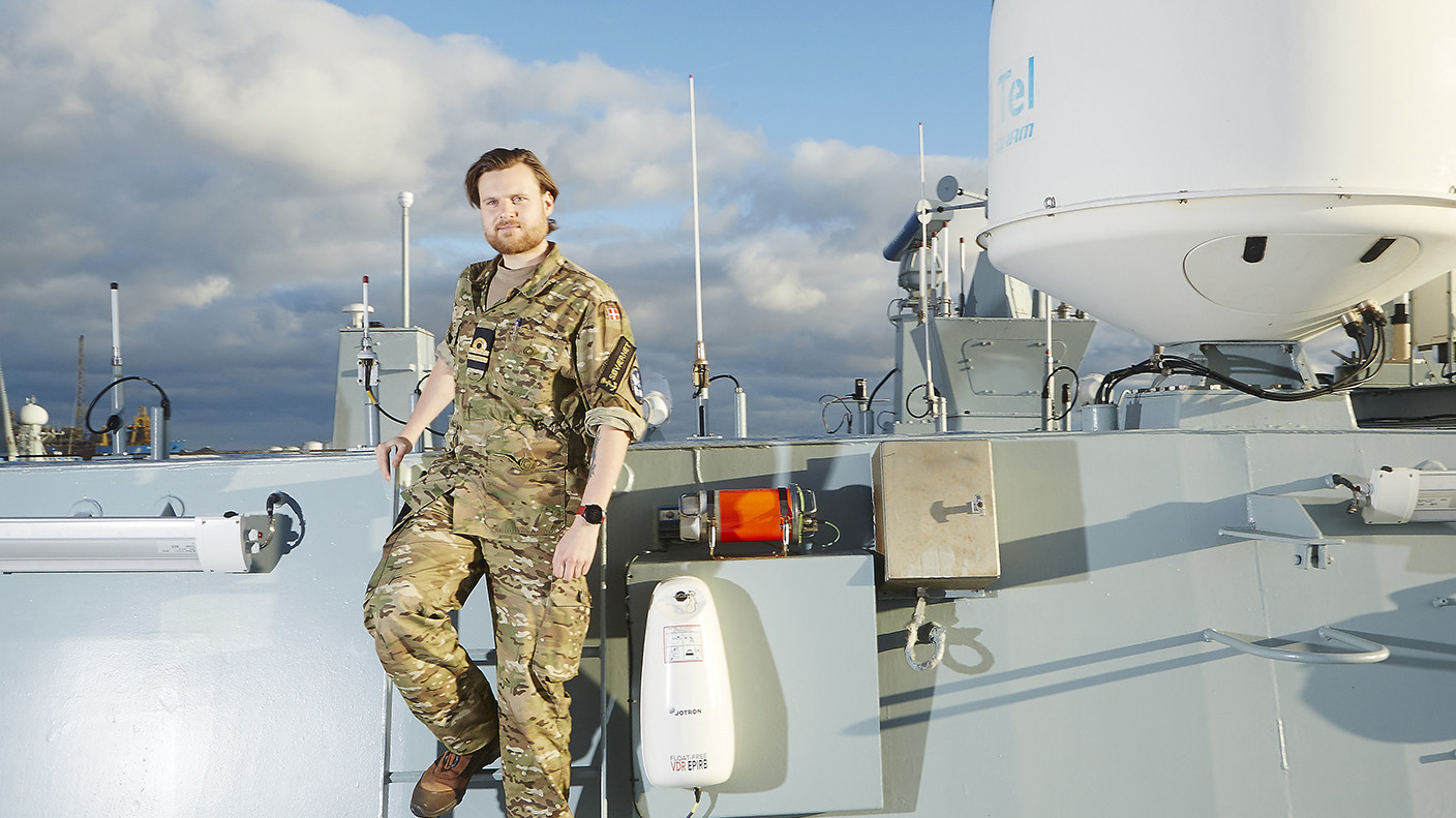 Mand i militæruniform med skæg står på et skib med kommunikationsudstyr og antenner i baggrunden under en overskyet himmel.